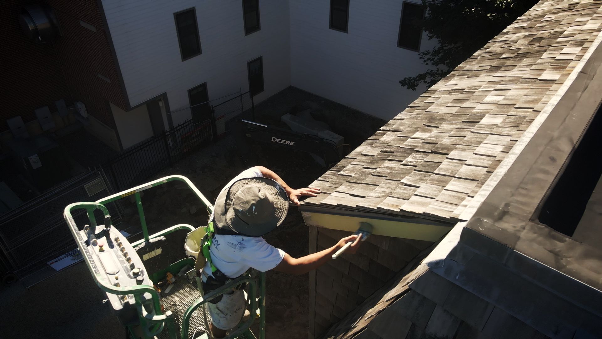 Person on a lift paints the edge of a building's roof. The painter is wearing a hat and harnesses, working outdoors.