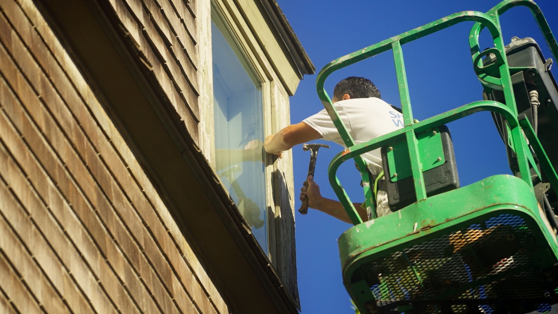 Person in a green lift hammering on a window frame of a wood-sided building under a blue sky.