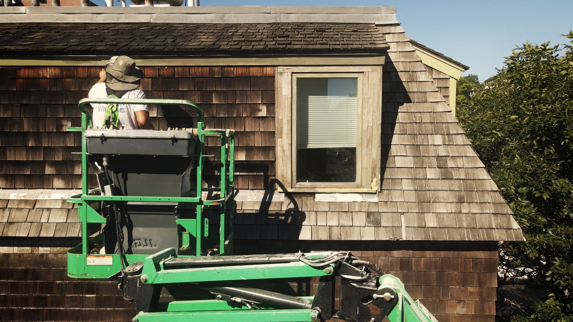 Person in a green lift working on a weathered, shingled house, near a window. Sunny day.