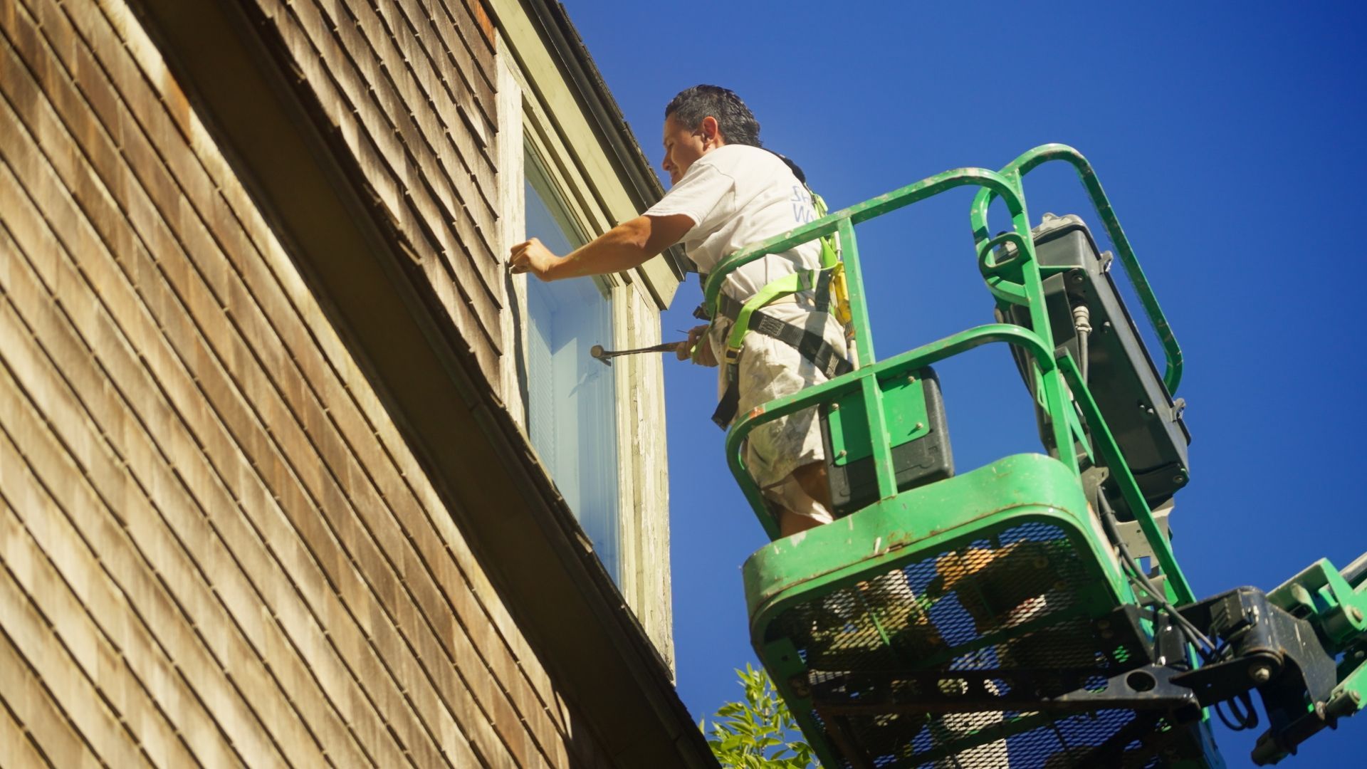 Man in a harness on a lift, cleaning a window of a wood-sided house on a sunny day.