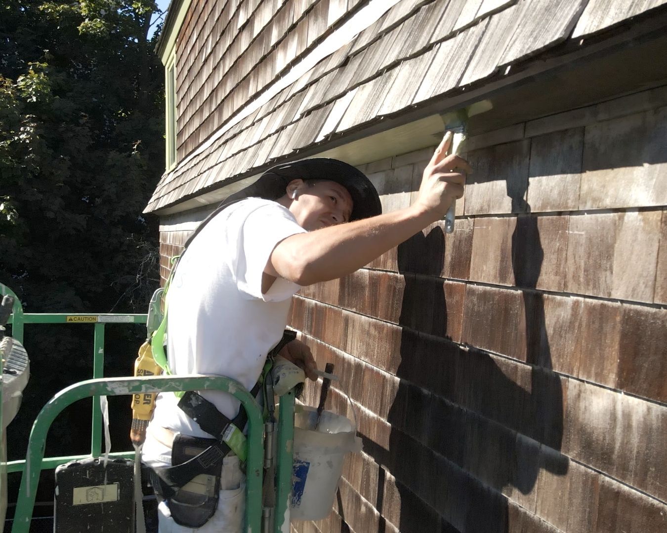 Man in a harness on a lift paints the trim of a weathered, wooden-shingled house.