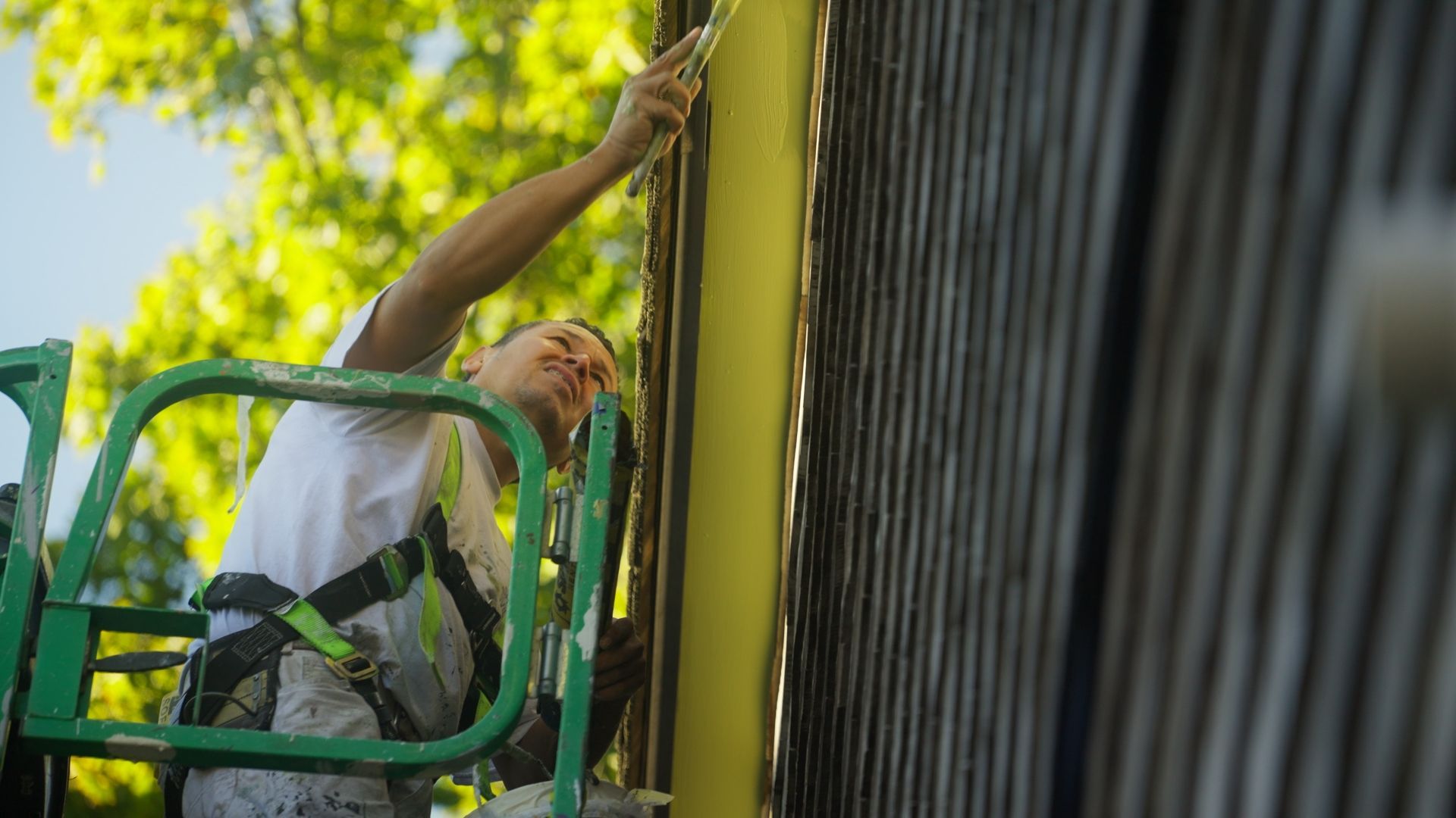 Man on a lift painting the exterior of a building yellow, wearing a harness. Green lift against a corrugated gray wall.