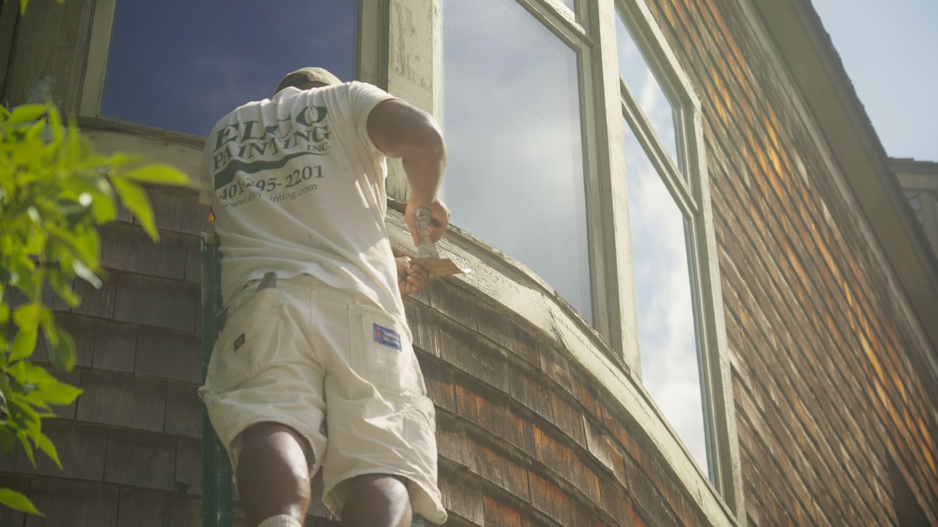 Man painting window trim on a shingled house, outdoors. Bright sunlight reflects off the glass.