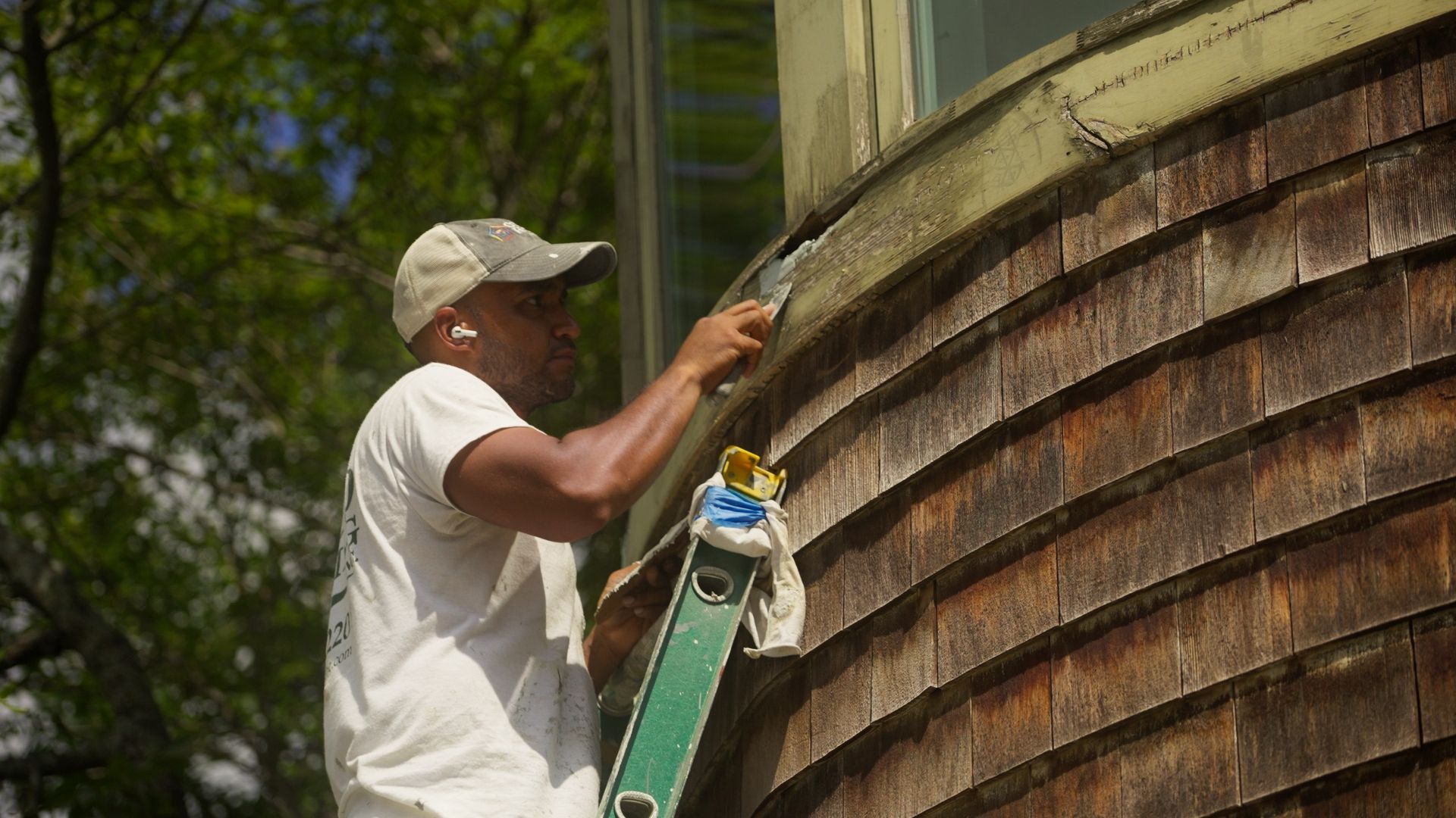 Man on ladder scraping weathered wood siding of a building. He wears a hat and white shirt, and the setting is outdoors on a sunny day.
