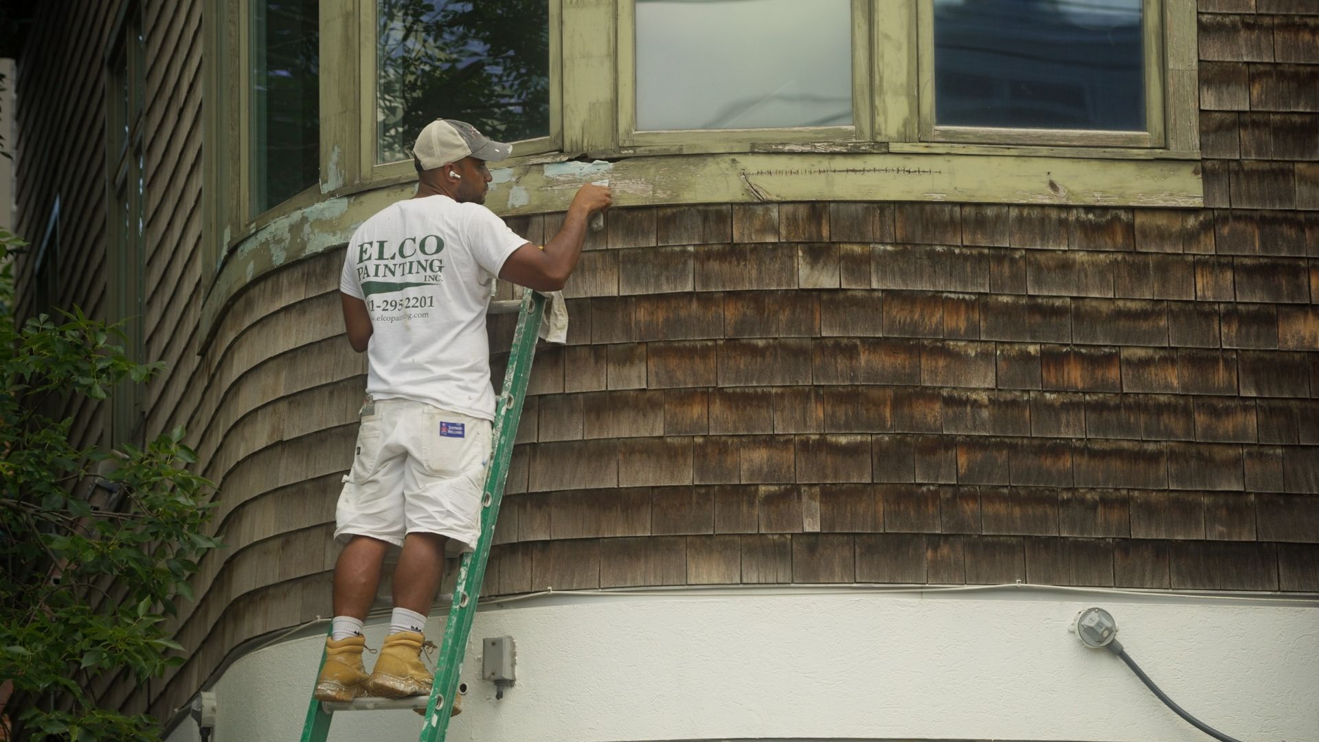 A person painting wood shingles on the side of a building while standing on a green ladder.