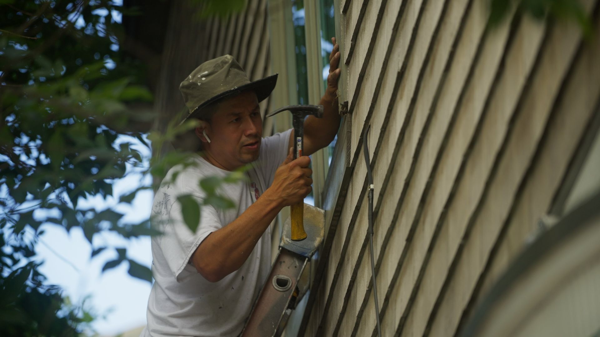 Man on a ladder hammering siding on a house; he wears a hat and white shirt.