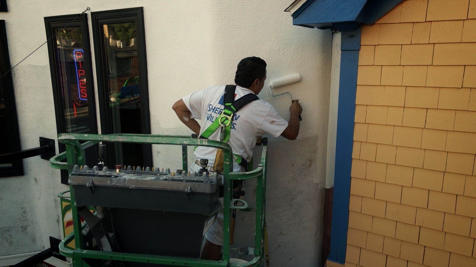 A painter in a lift rolls white paint onto a stucco building. The building is partially yellow and has a blue overhang.