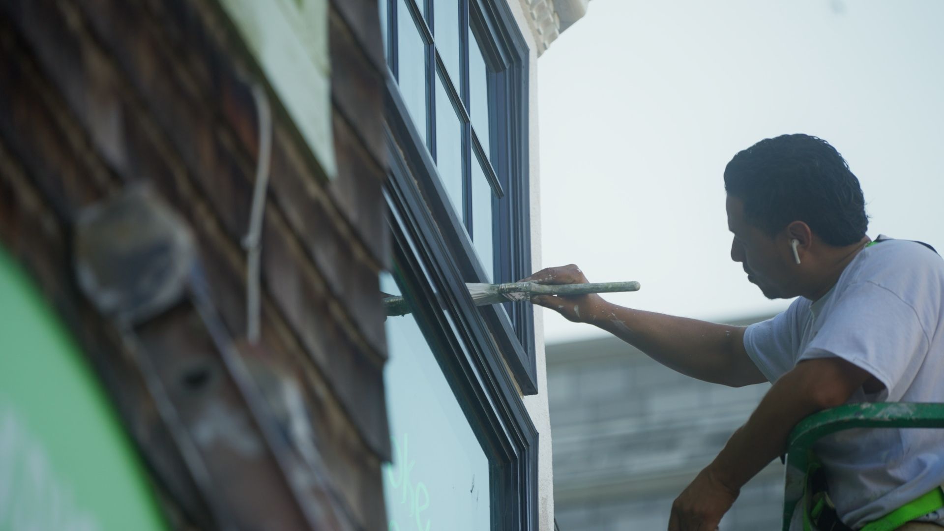 A man painting a window frame on a building exterior, using a brush. He is wearing a light-colored shirt and appears to be working from a lift.