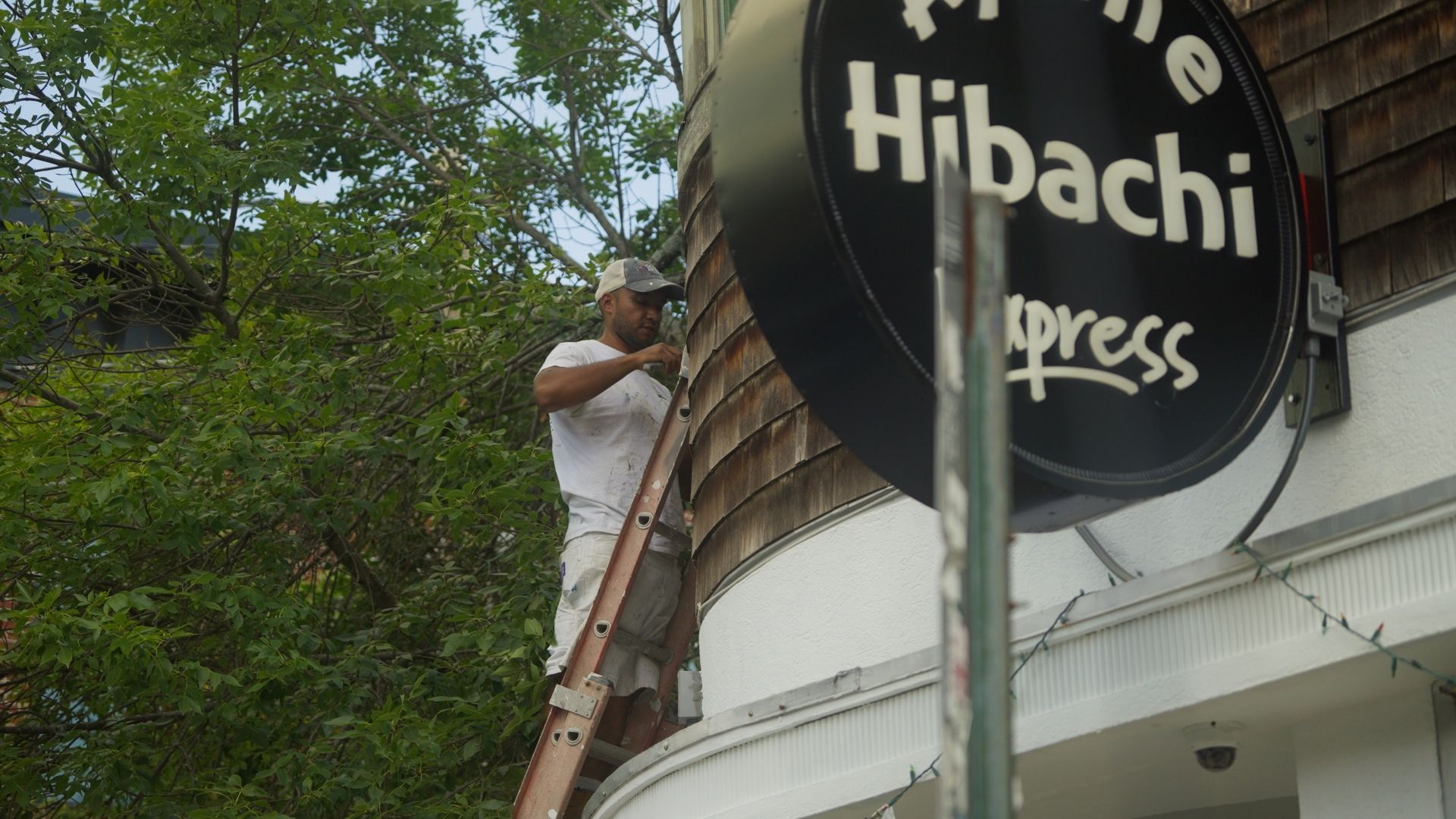 A man on a ladder repairs the wooden siding of a building with a large, round