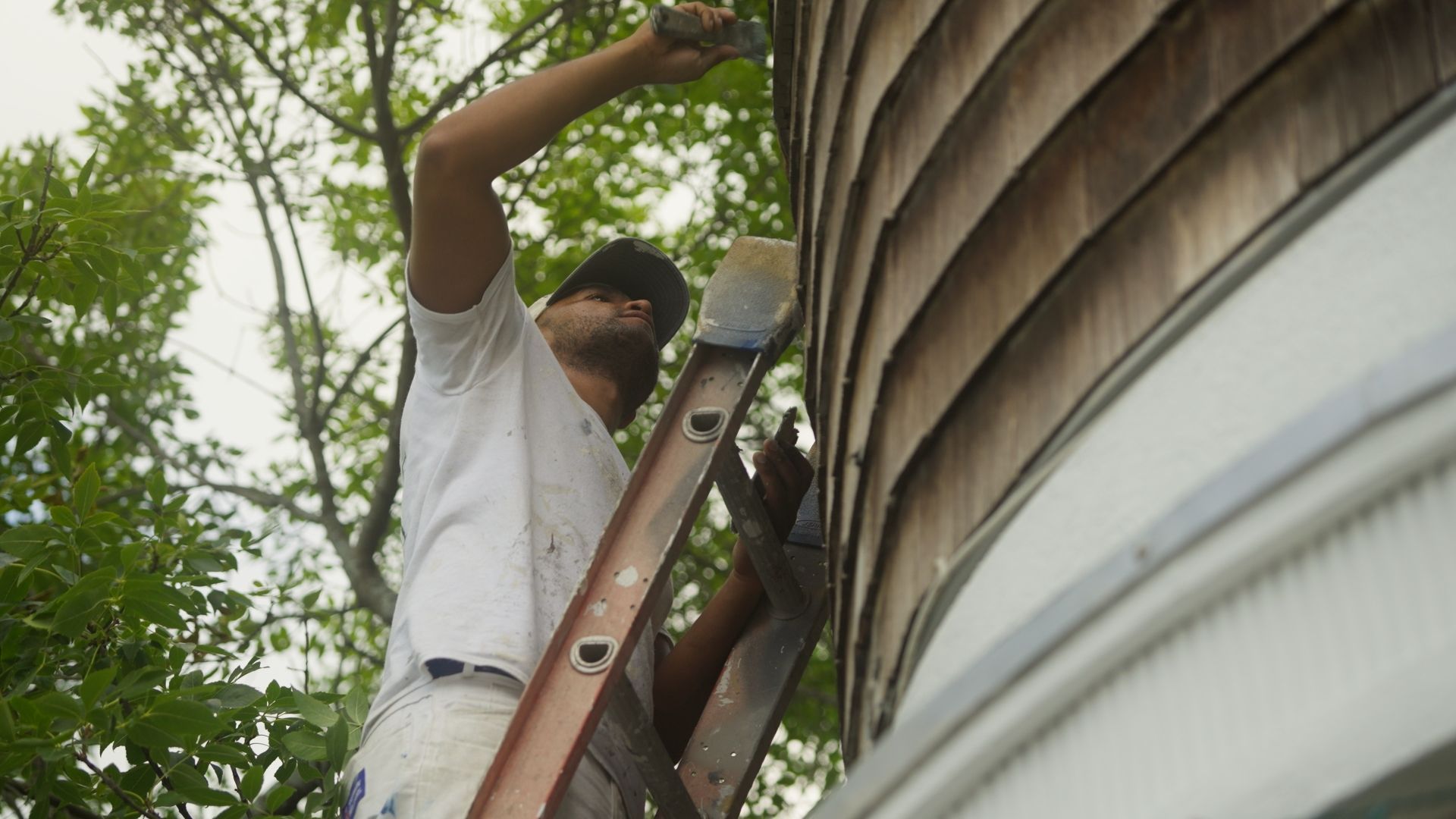 Man on a ladder painting the side of a building with a brush.