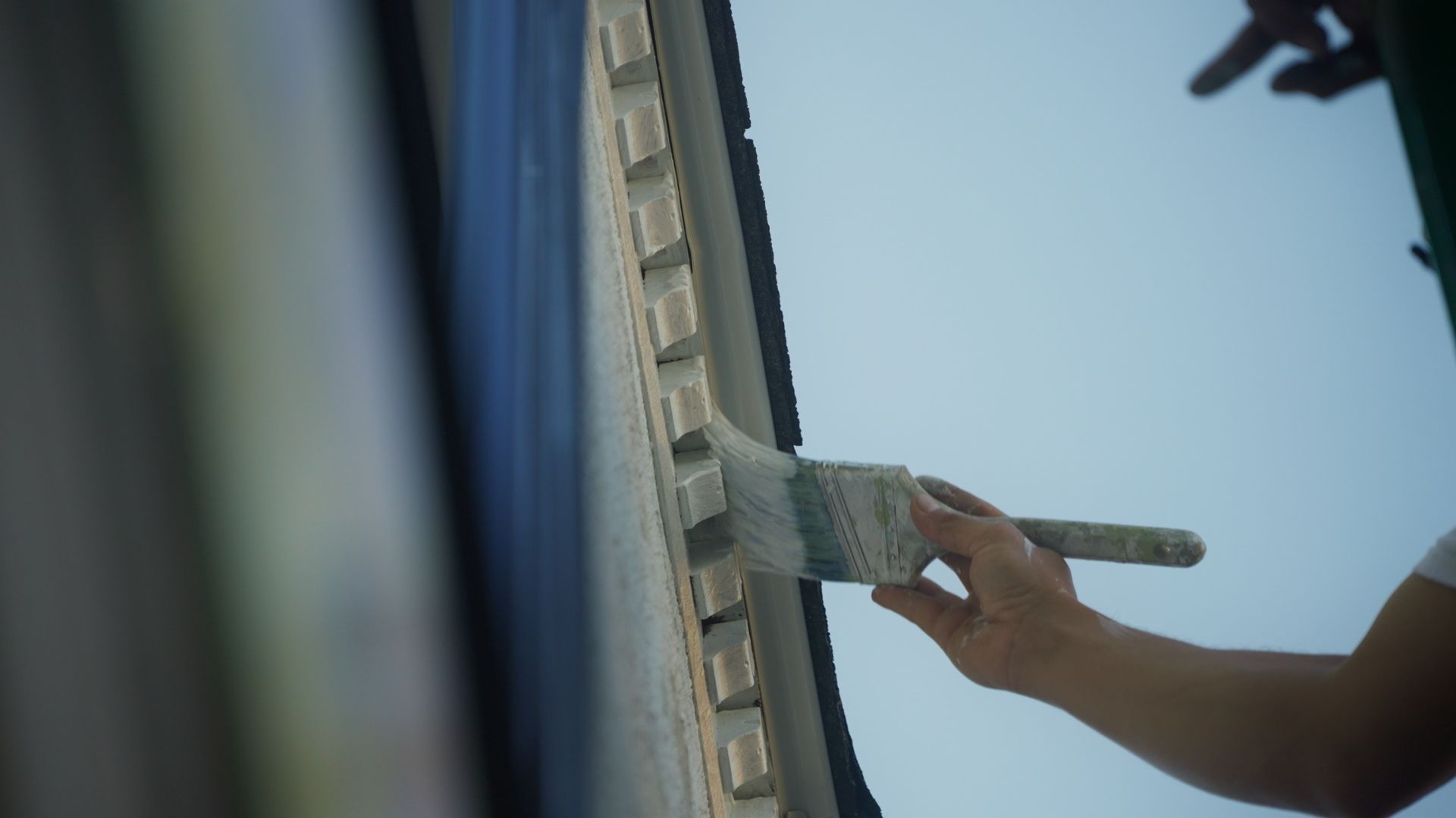 A person paints a light-colored trim on the side of a building with a brush, against a blue sky.