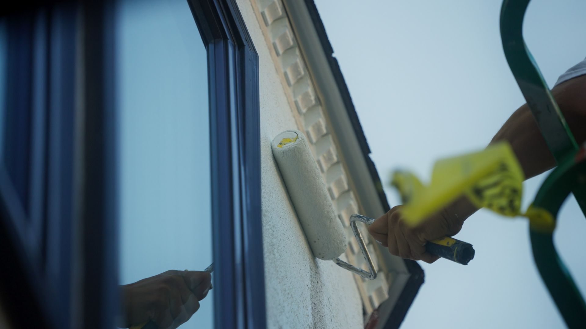 Person painting the exterior of a house with a paint roller; a ladder and window are also visible.