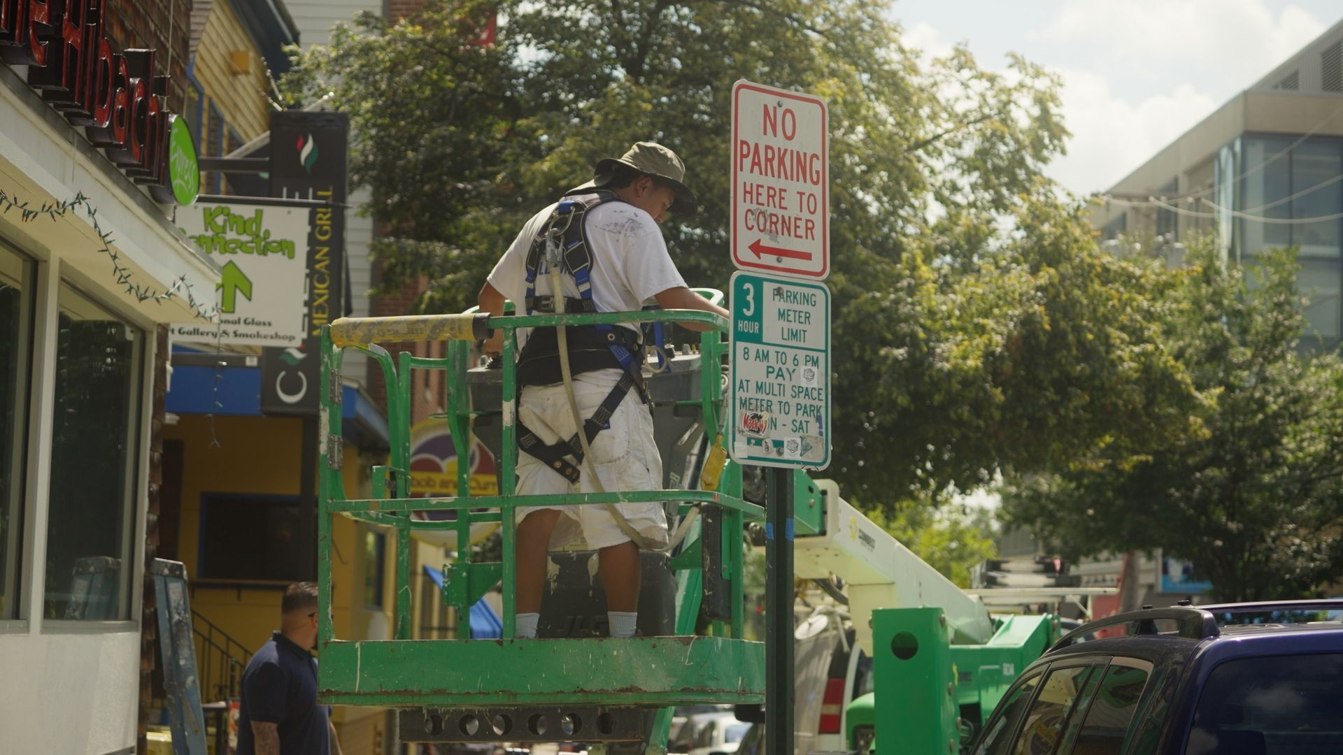 Man in a harness on a lift is working on a street sign. He's outside near a building and cars.