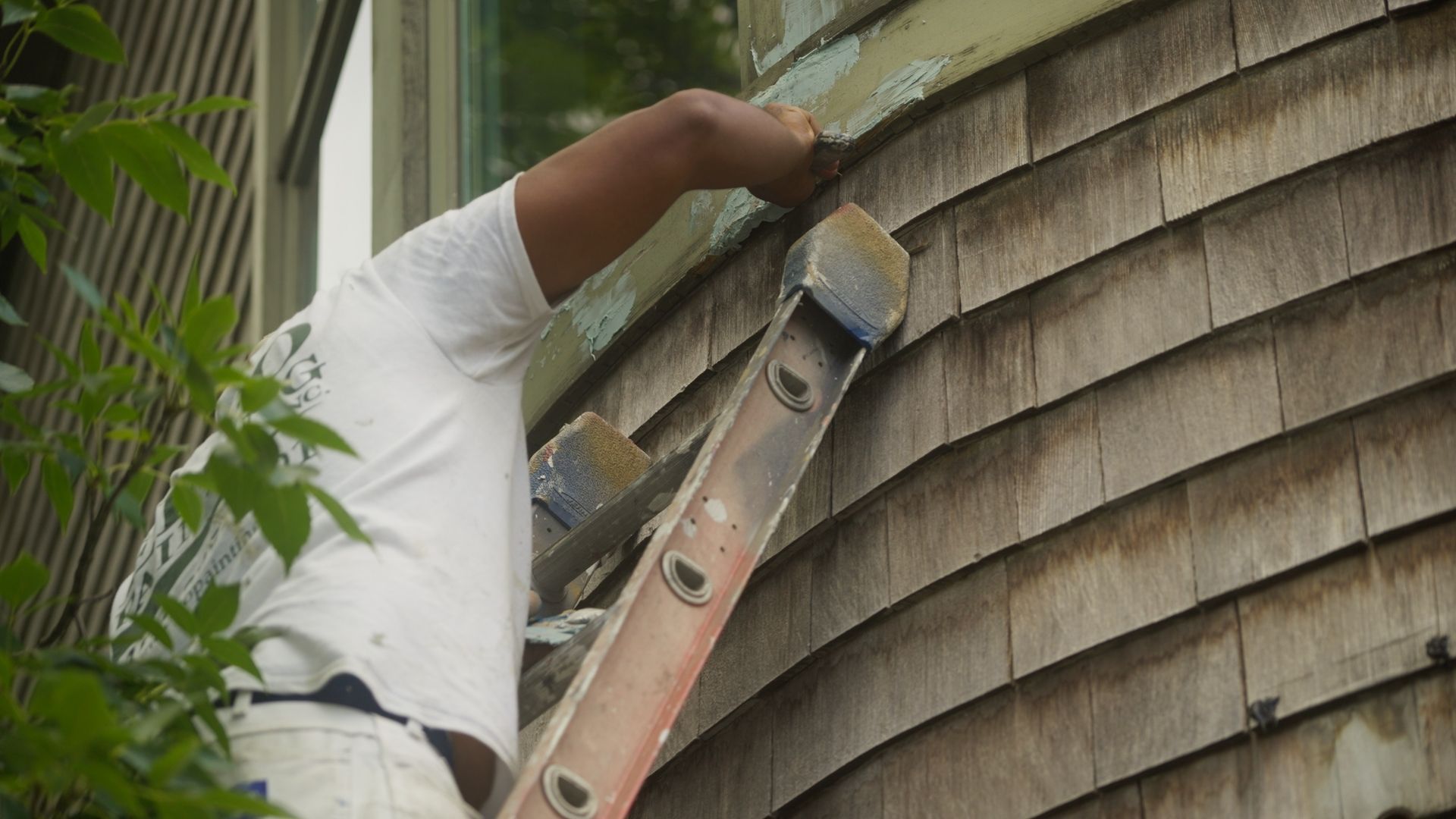 Person on a ladder scraping paint from the side of a wooden shingled house.