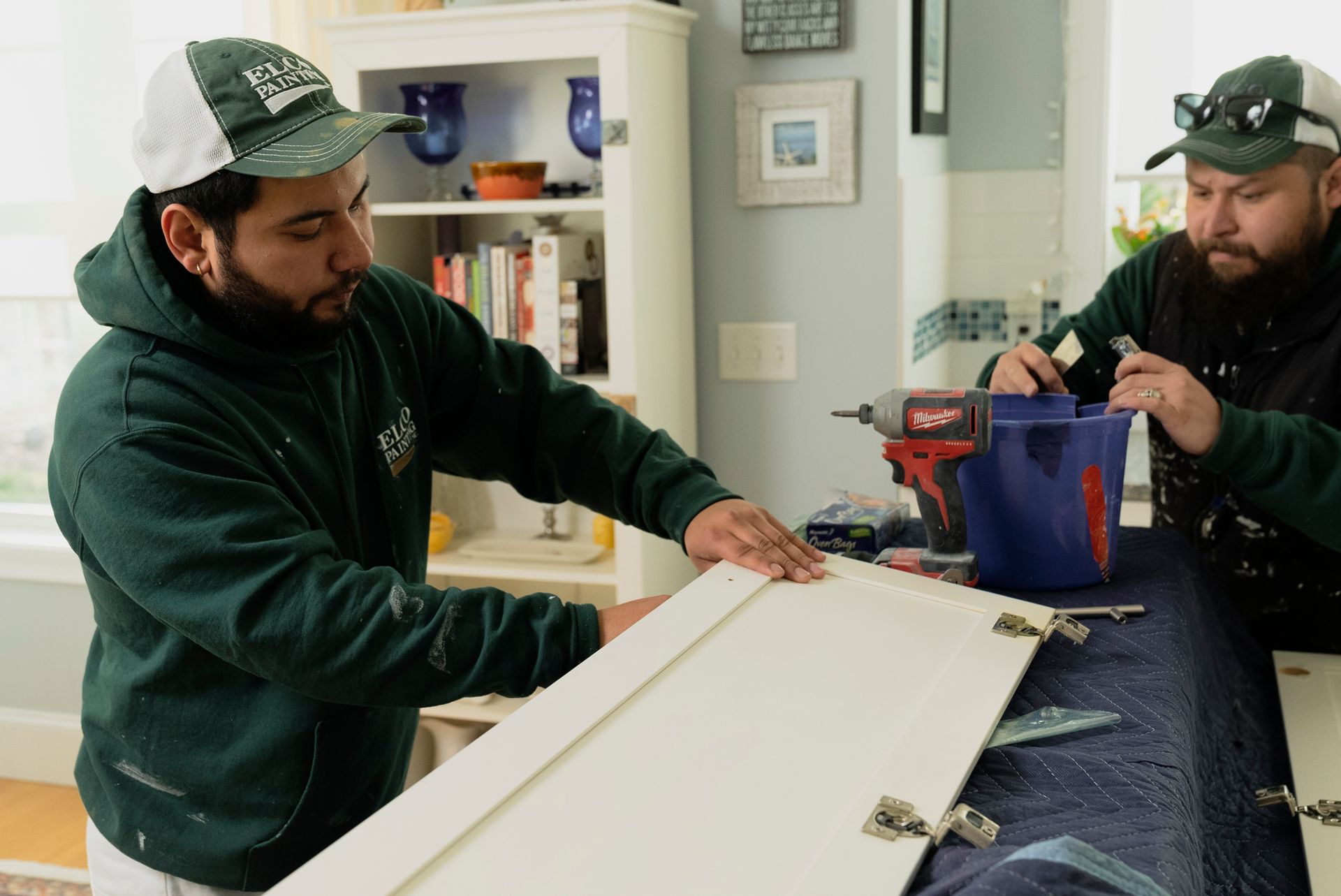 Two men installing a white cabinet door in a kitchen. One uses a drill while the other looks on, both wearing green shirts and caps.