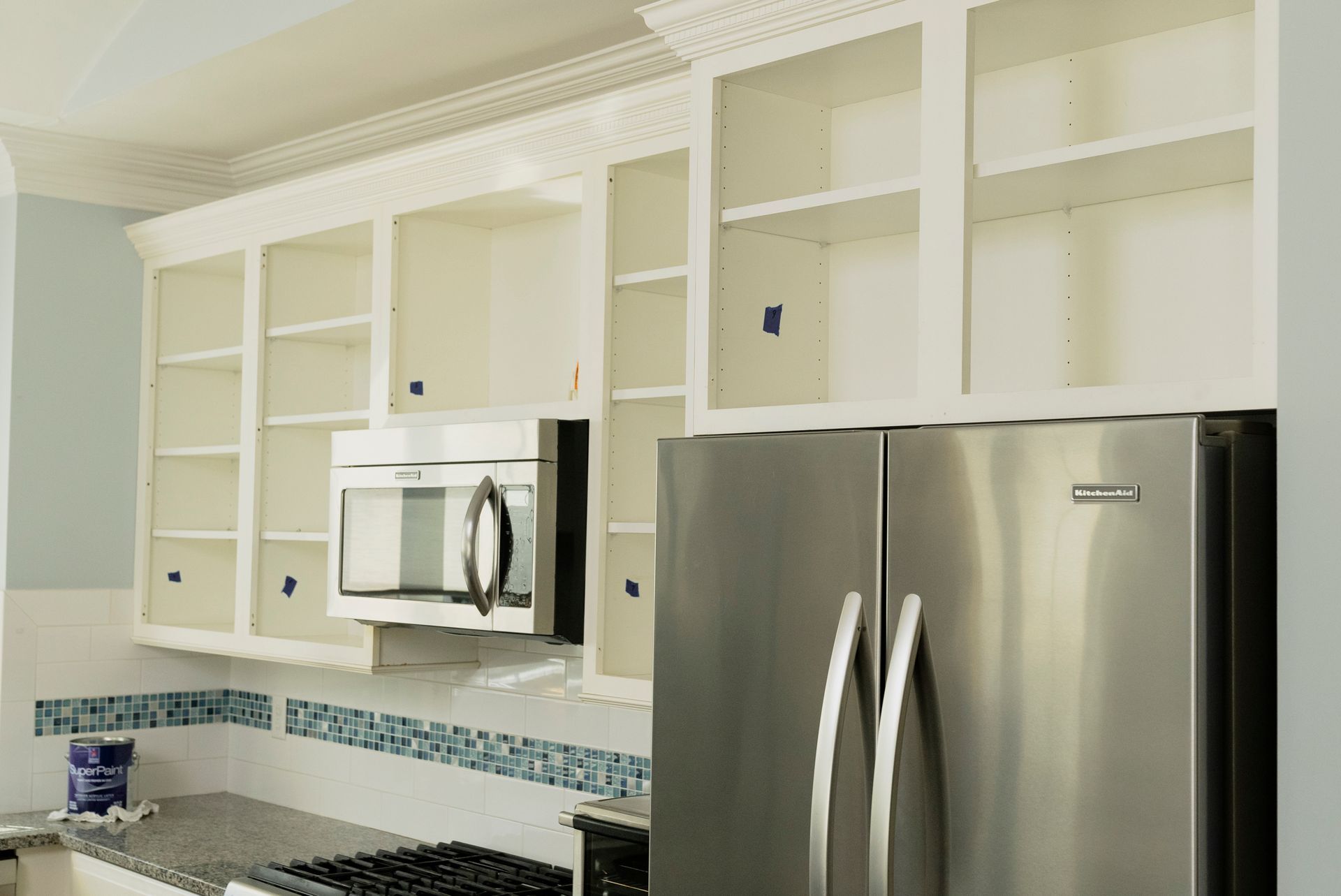Empty white kitchen cabinets above a stainless steel refrigerator and microwave. Light blue backsplash visible.