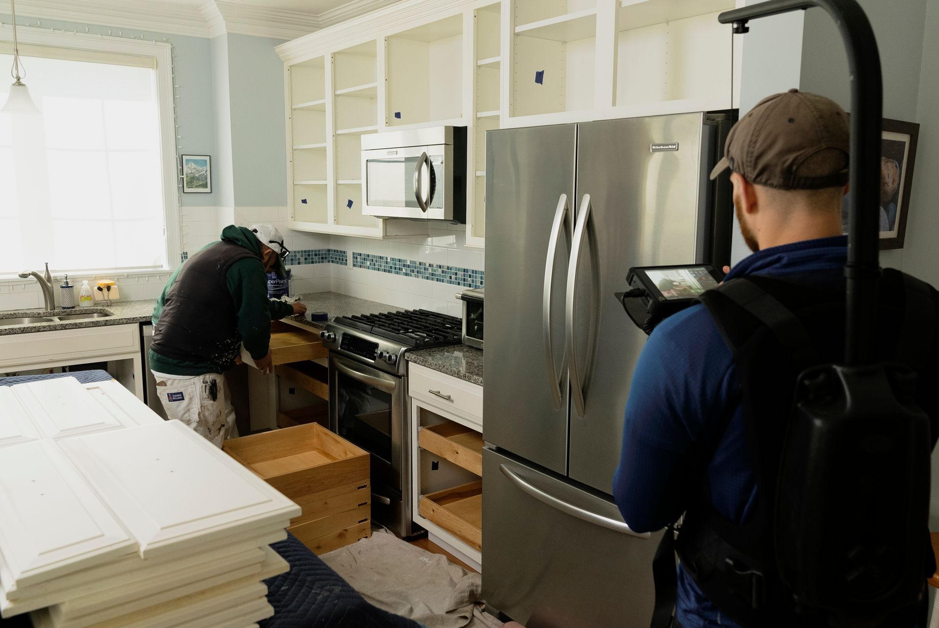 A kitchen renovation in progress. Two men work on cabinets and appliances, one filming the project with a stabilizing camera rig.