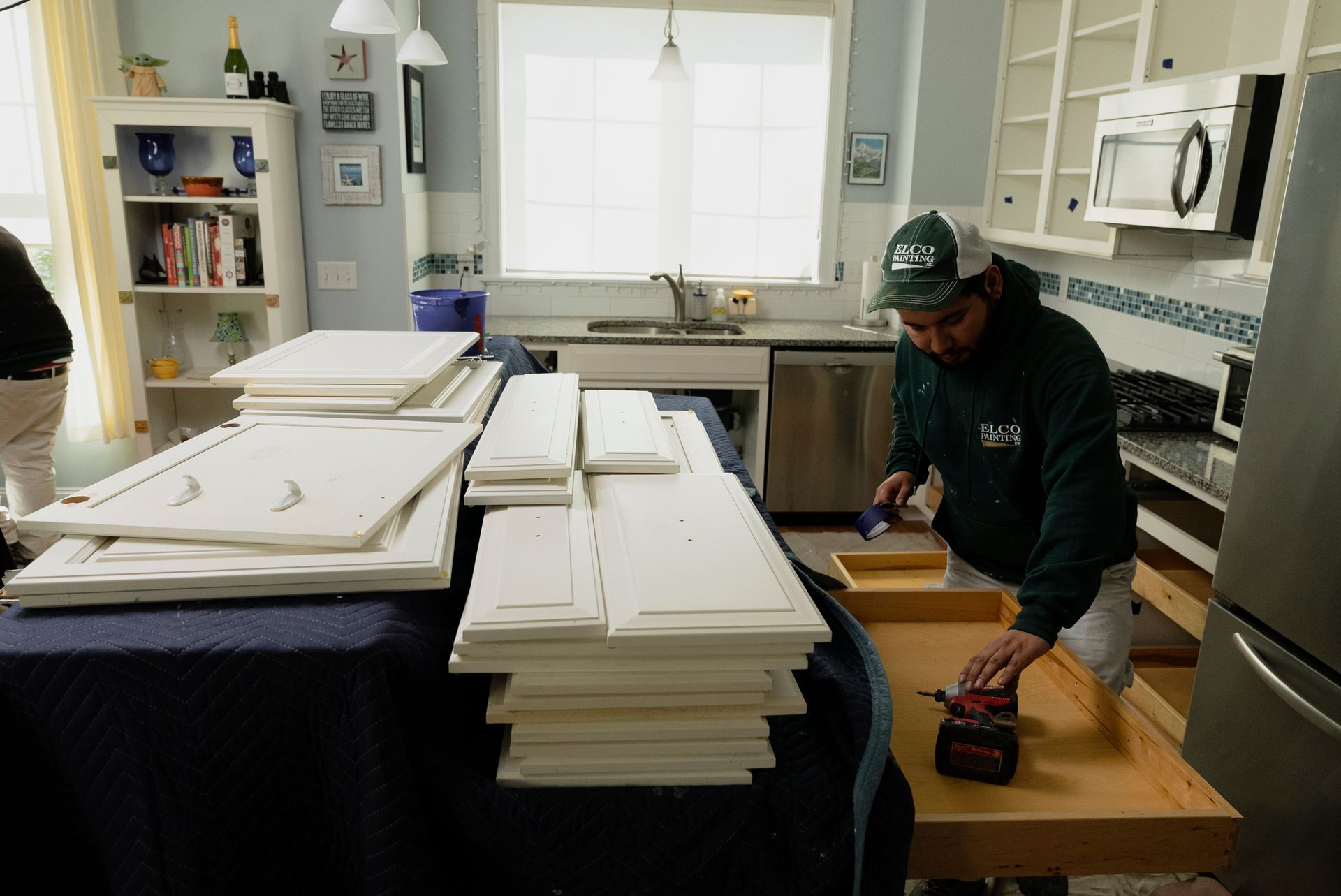 Man assembling cabinet doors in a kitchen; piles of white cabinet pieces on a table.