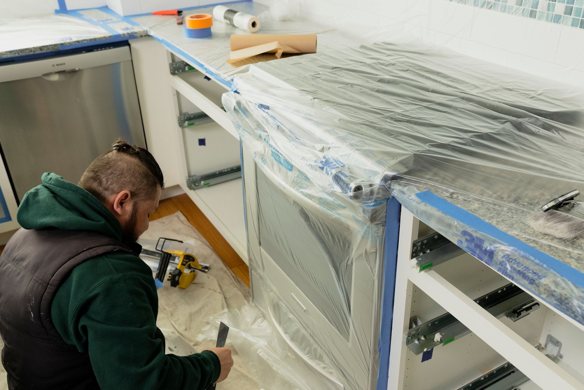 A person wearing a vest taping and covering kitchen cabinets with plastic sheeting in a renovation.