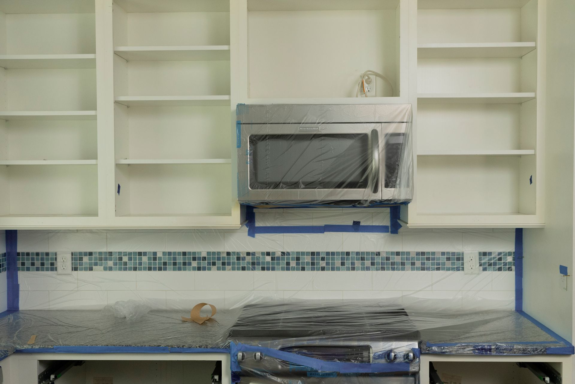 Kitchen with upper cabinets, microwave, and tiled backsplash. The stove and countertop are covered in plastic for protection.