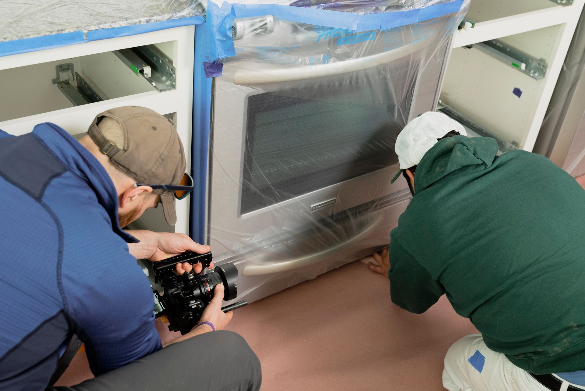 Two men in a kitchen, one filming the other who is kneeling near a plastic-covered oven. Cabinets are also protected with plastic.