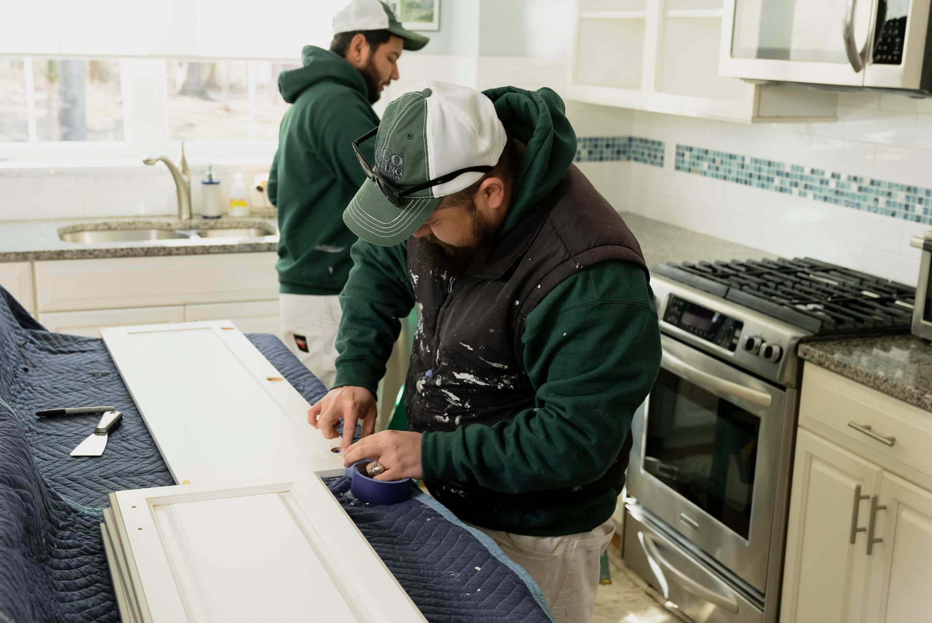 Two men painting kitchen cabinets white in a kitchen. One is working on a cabinet door on a drop cloth.