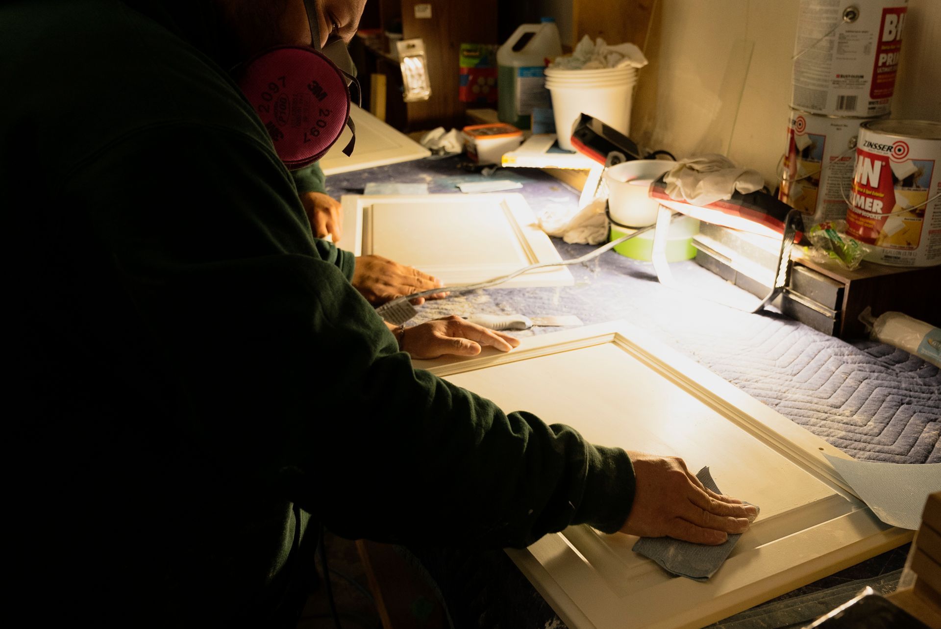 Person wearing a respirator sanding a white cabinet door in a workshop. A second door rests on the work surface.