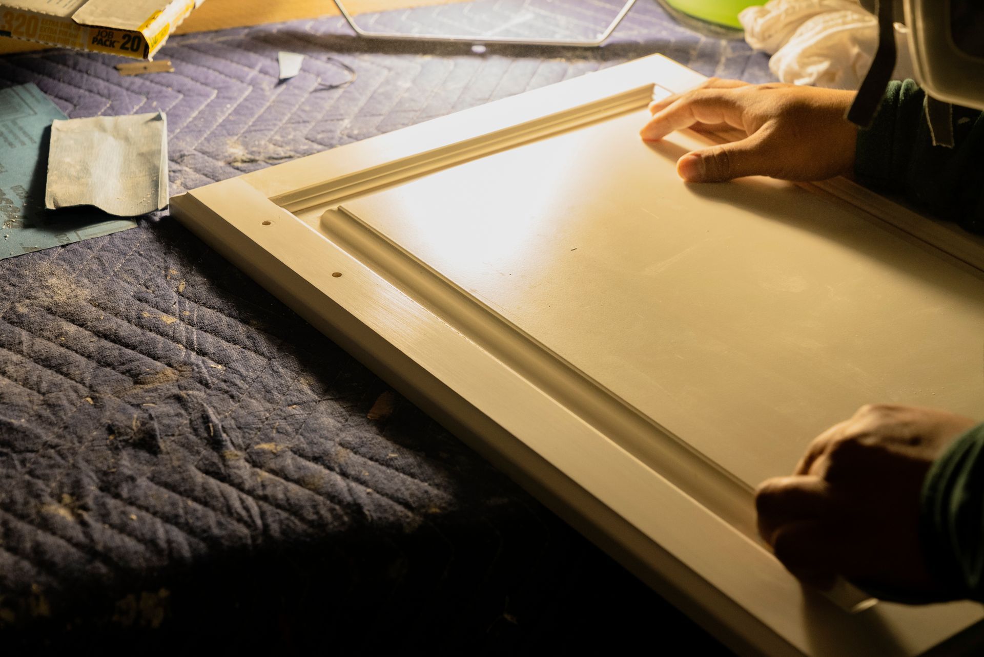Hands inspect a freshly painted white cabinet door resting on a blue surface. Sandpaper is visible nearby.