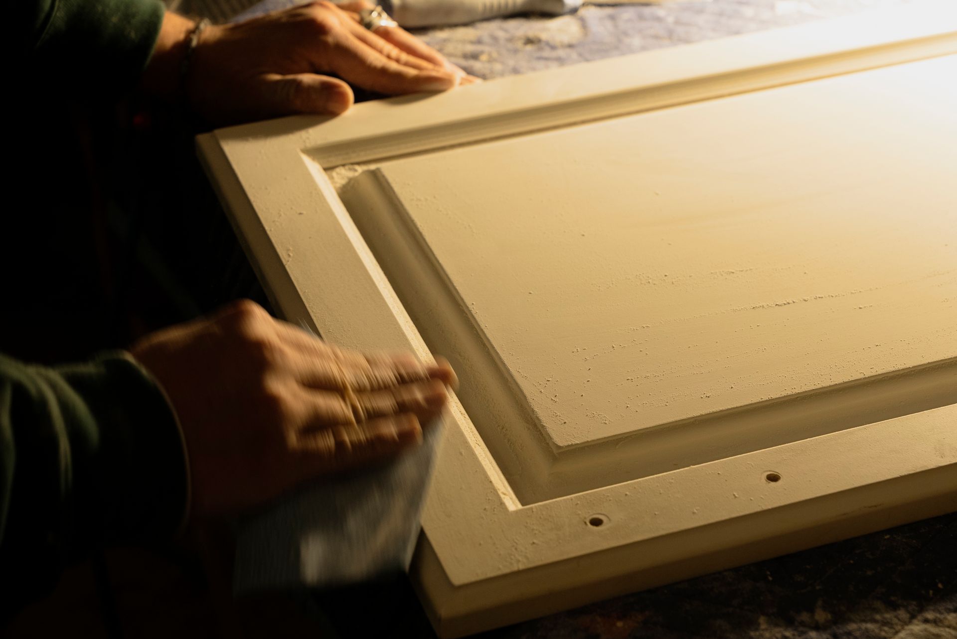Person sanding a white cabinet door. Hands are holding sandpaper, smoothing the frame.