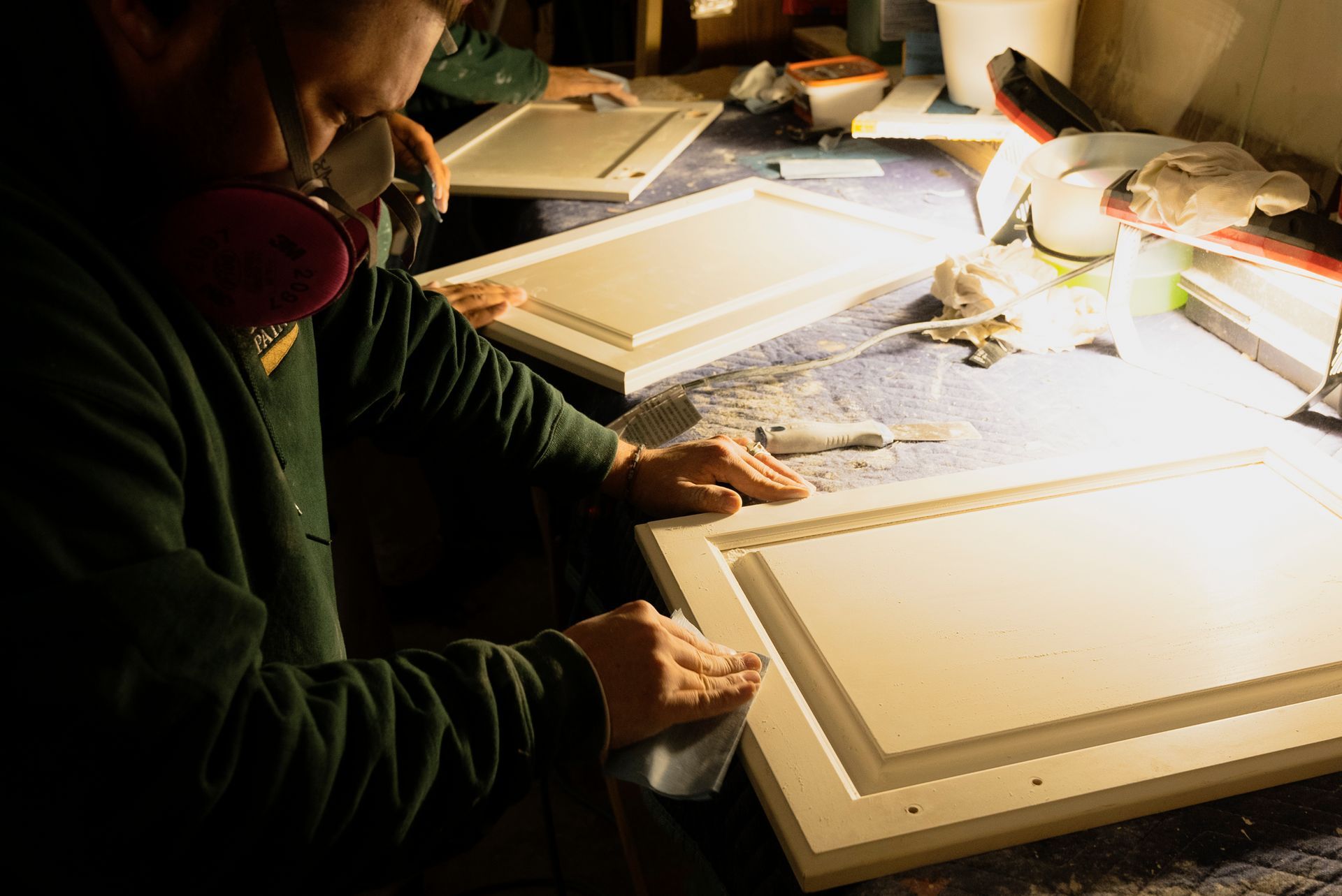 Person sanding a white cabinet door while wearing a respirator in a workshop.