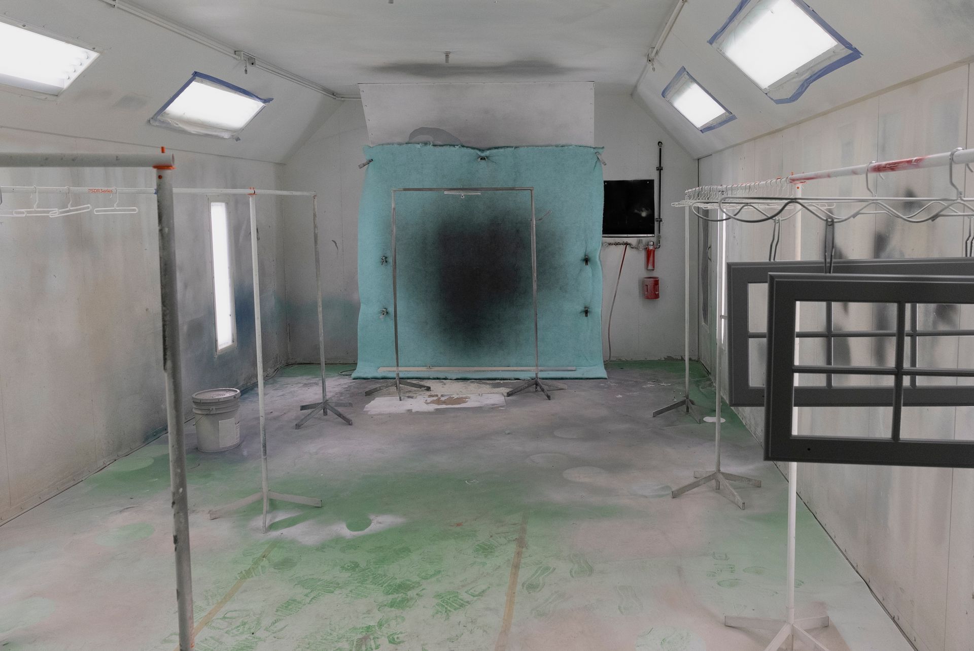An empty spray booth with a green floor and a blue backdrop shows dark paint marks, along with window frames hanging on stands.