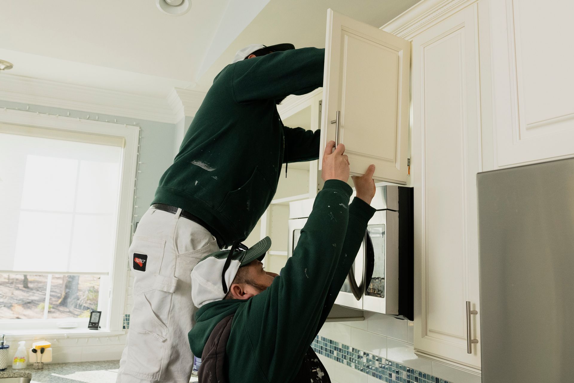 Two workers in a kitchen, one standing on a ladder, installing a white cabinet door.