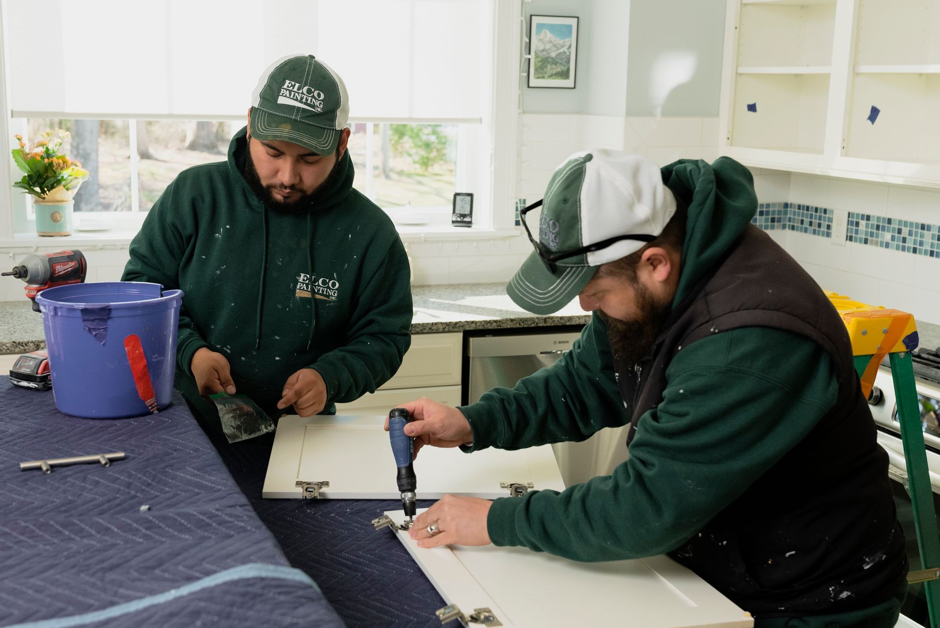 Two workers in green hoodies install cabinet doors in a kitchen. One uses a screwdriver; the other scrapes.