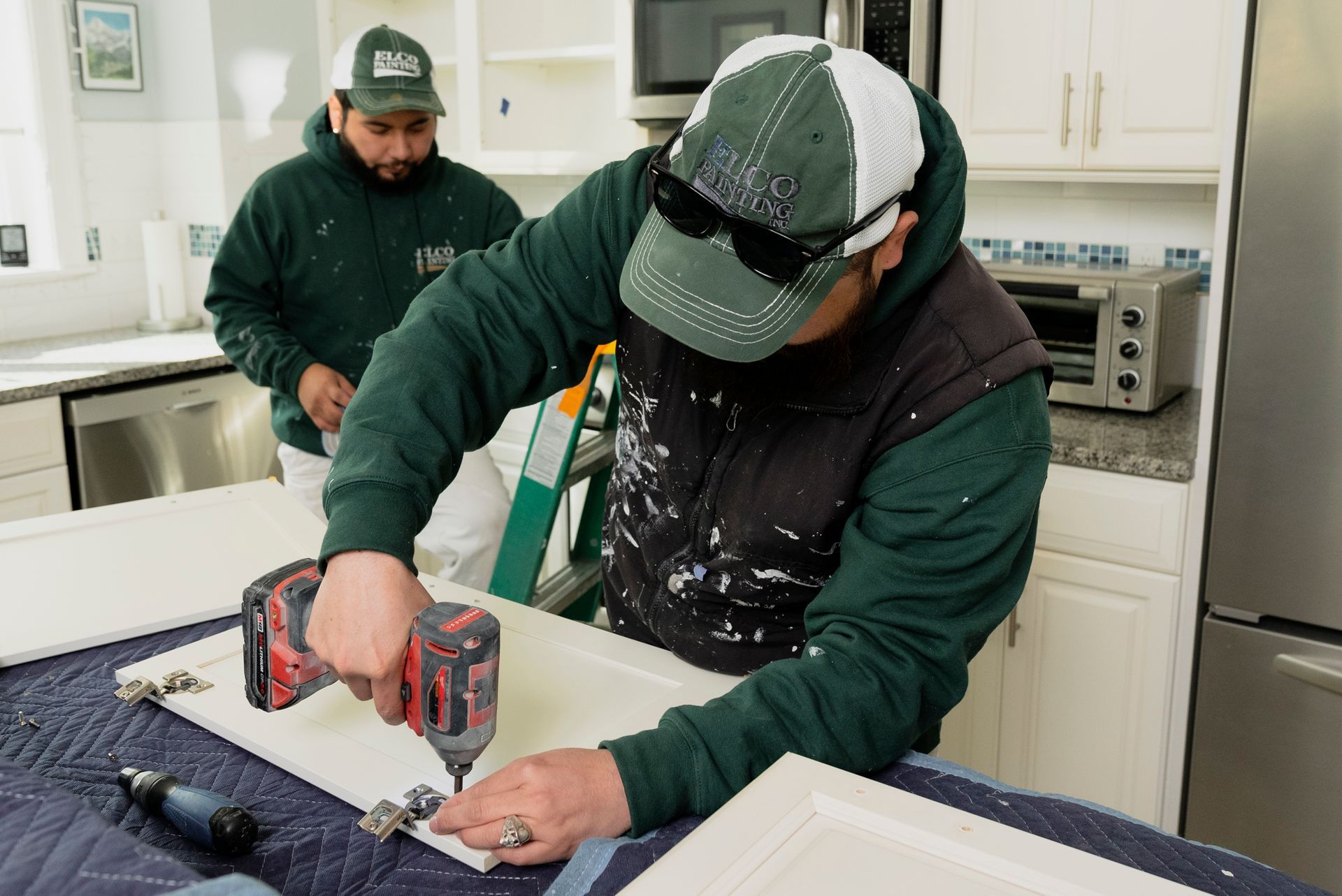Two men in a kitchen installing cabinet doors. One uses a drill, the other watches.