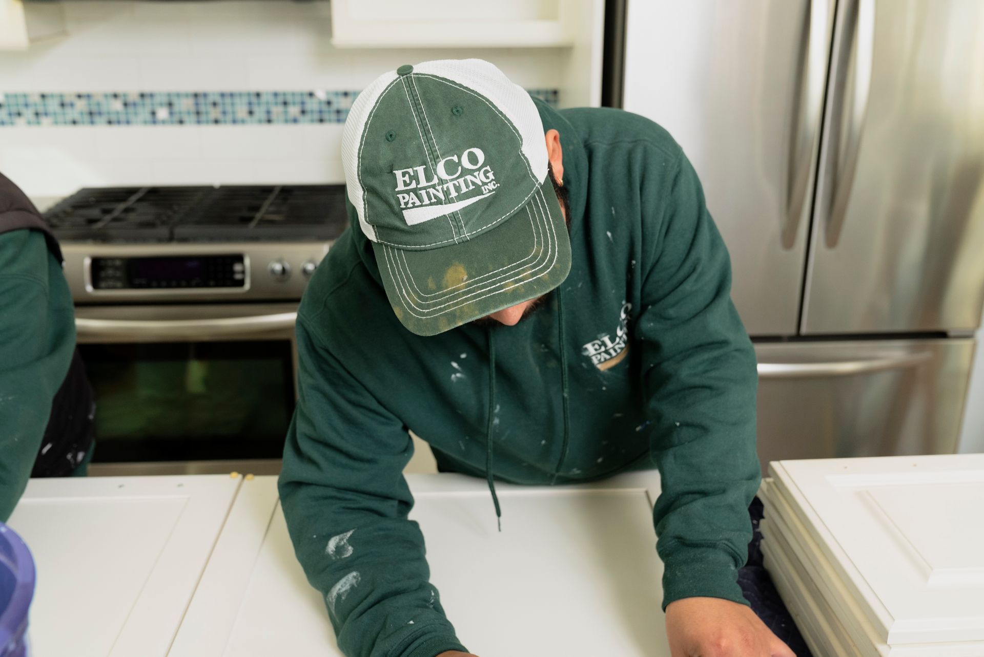 A person wearing a green sweatshirt and a hat is leaning over white cabinet doors in a kitchen.
