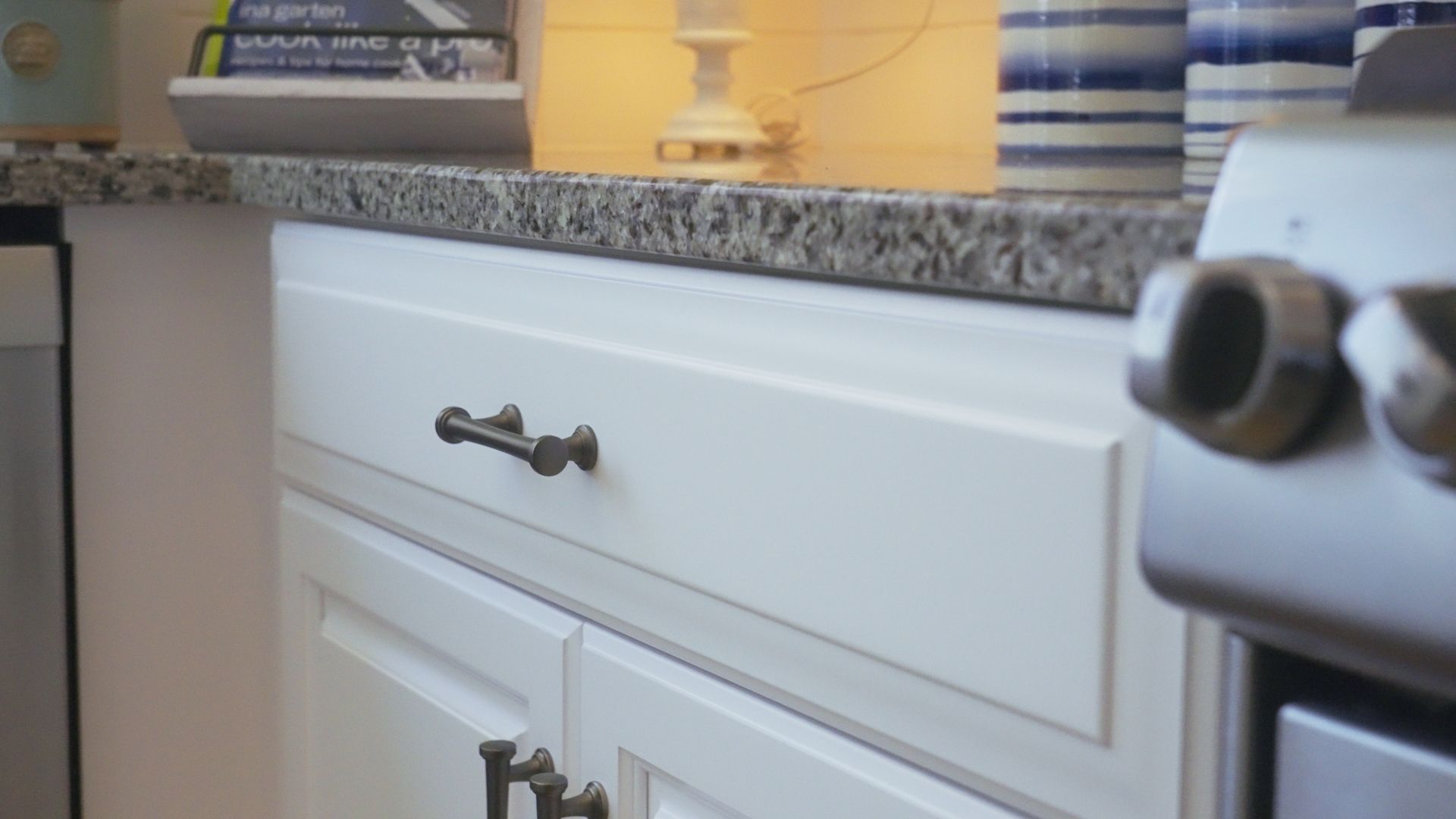 White kitchen cabinets with dark metal hardware under a granite countertop, next to a stainless steel stove.