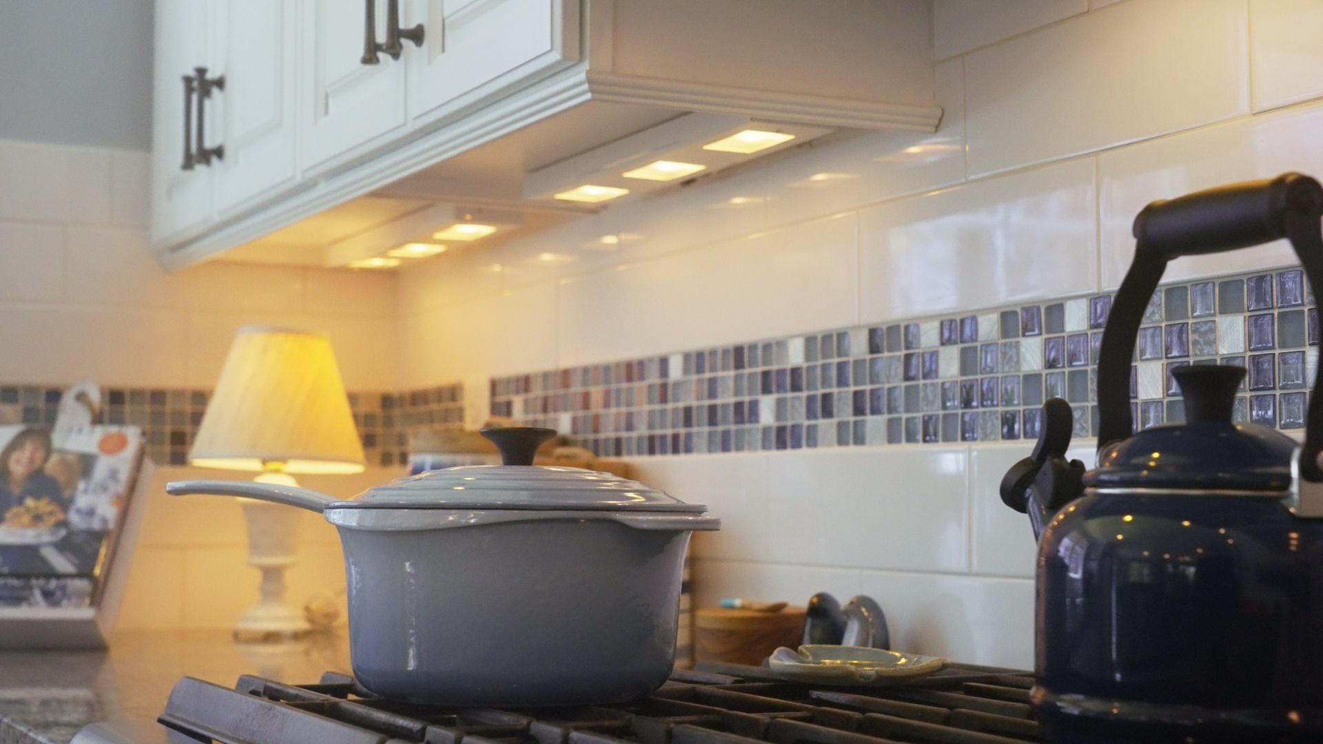 Kitchen scene with a lit stove, pot, kettle, and under-cabinet lights. Tile backsplash in a blue and white mosaic.