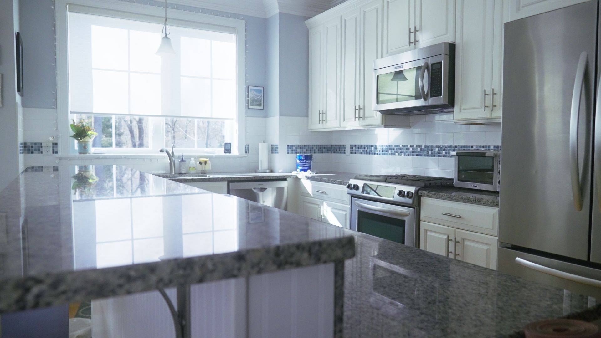 A bright, well-lit kitchen with white cabinets, stainless steel appliances, and a granite countertop. Sunlight streams in from a window.