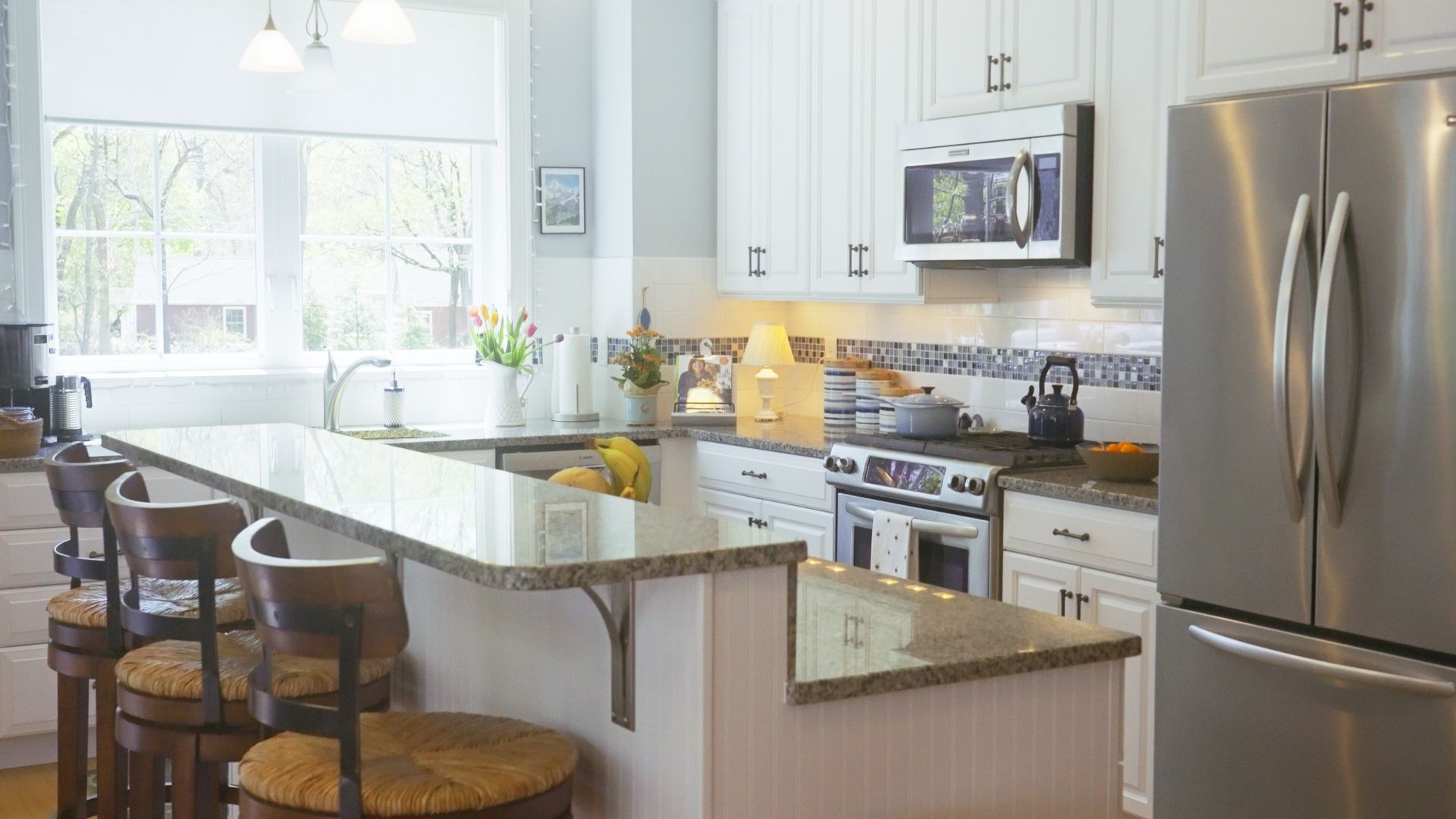 A bright kitchen with a granite countertop island, white cabinets, and stainless steel appliances. Bar stools are at the counter.