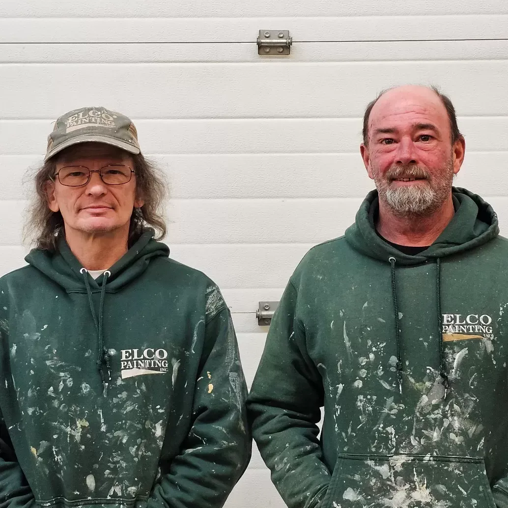 Two men wearing paint-splattered green hoodies stand in front of a white garage door.
