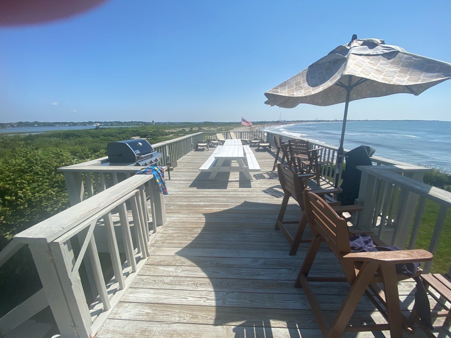 A long wooden walkway on a beach with a picnic table, chairs, grill, and umbrella. The ocean is on one side, and bushes are on the other, under a bright blue sky.