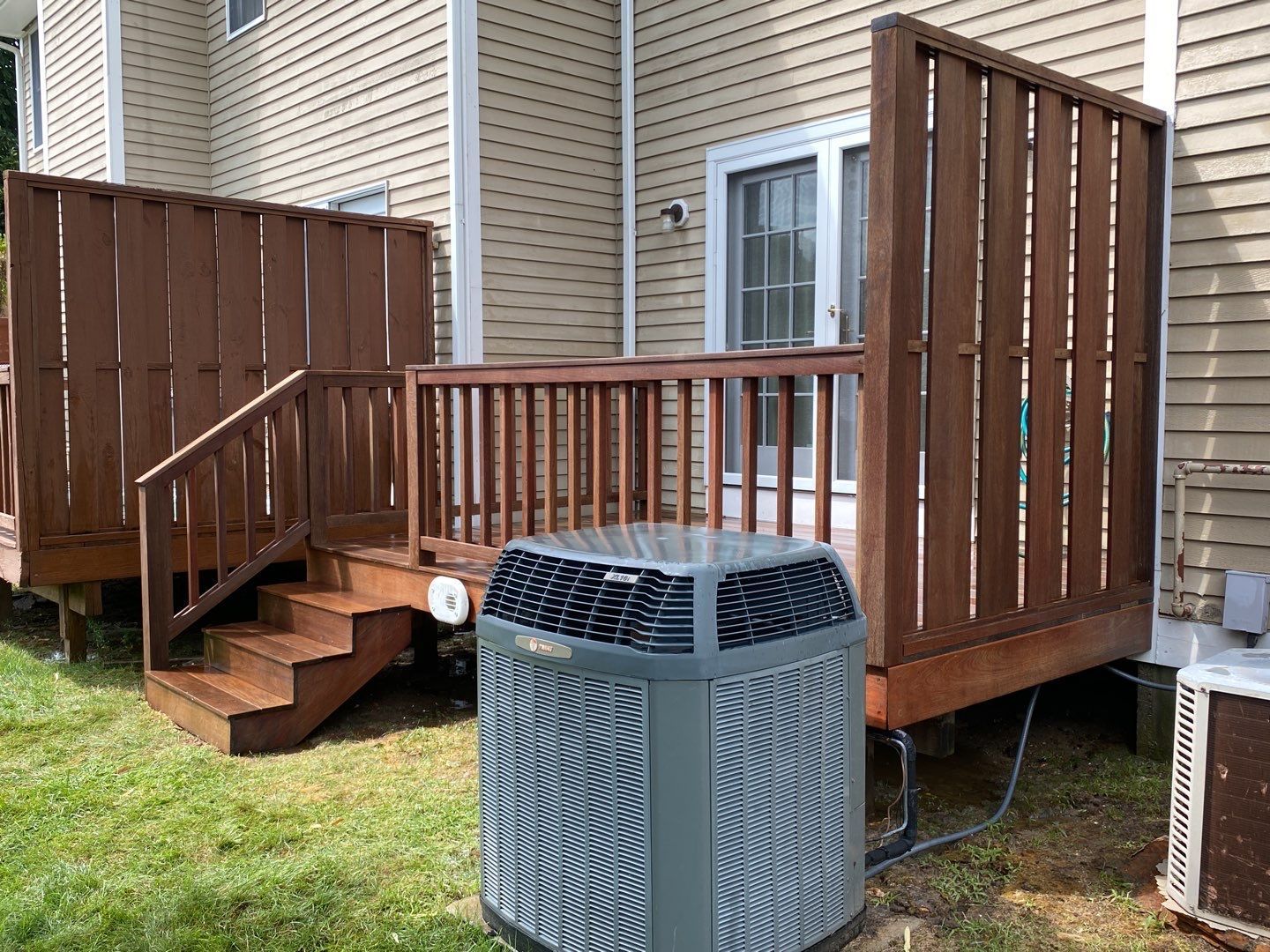 A deck with brown wooden railing and steps, a gray air conditioning unit in front. The building is beige with a sliding glass door.
