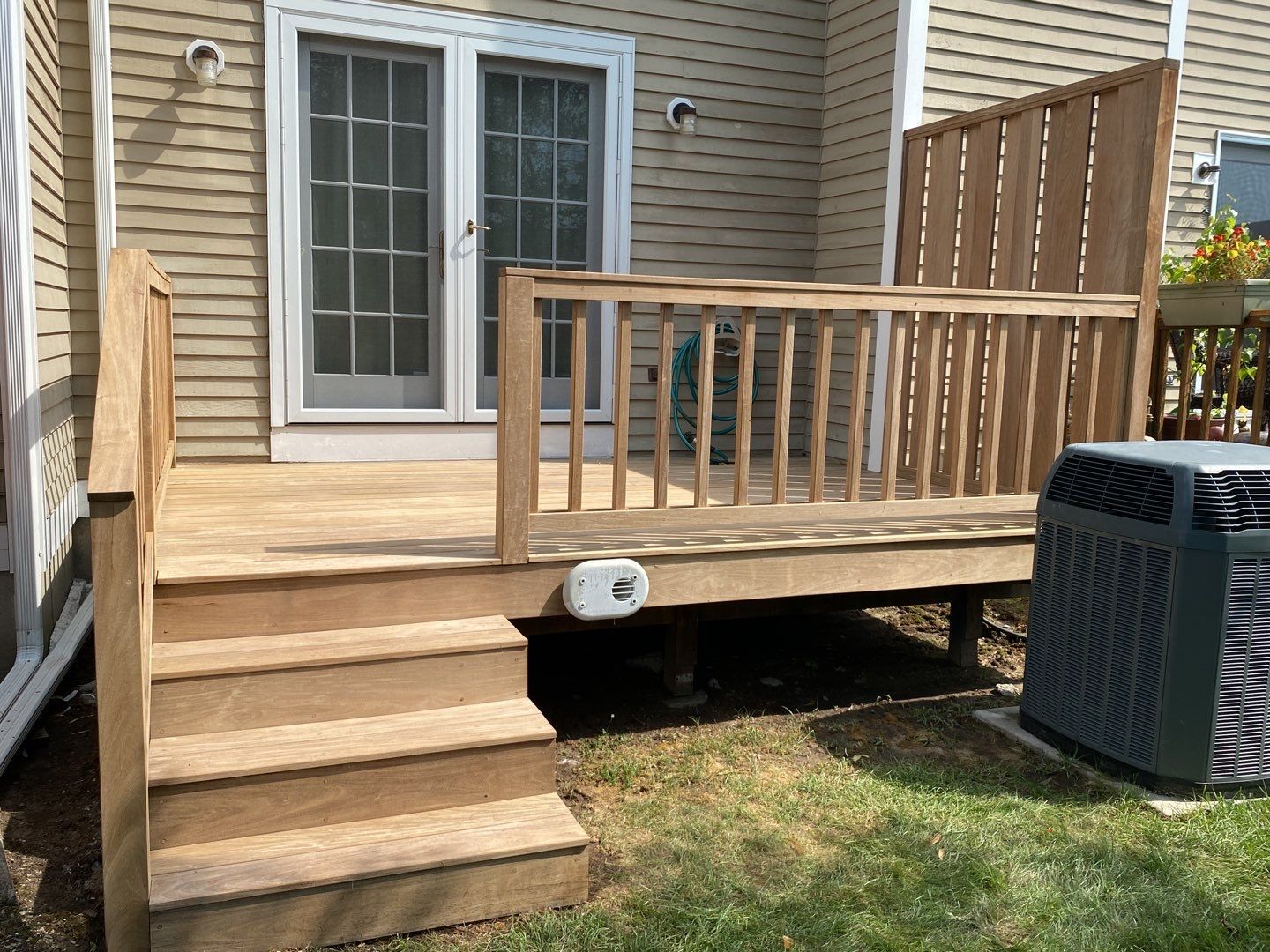 Wooden deck with stairs leading to a set of double doors, railing, and an air conditioning unit on a lawn.