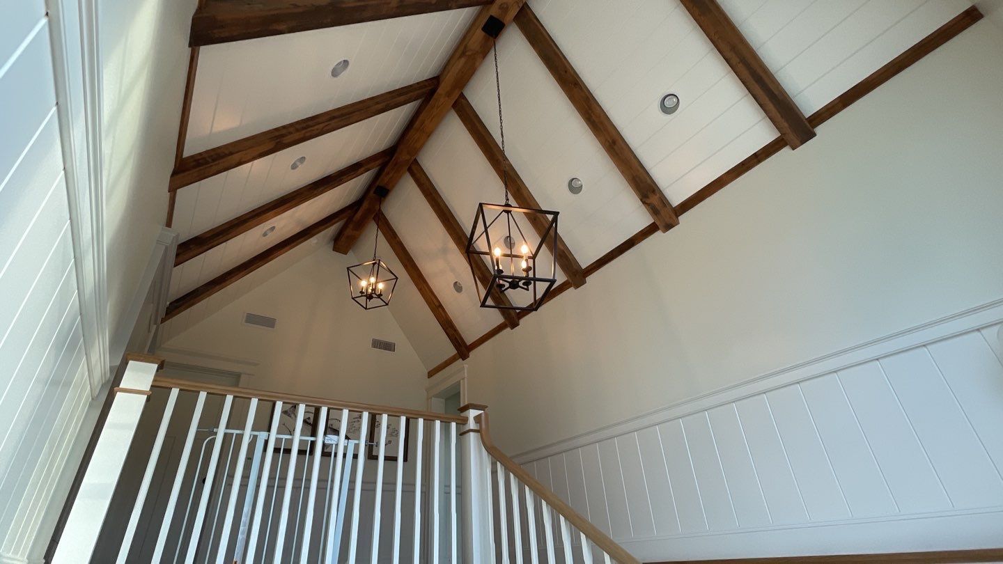 High-angle view of a white vaulted ceiling with exposed wooden beams and two pendant lights. A staircase railing is visible below.
