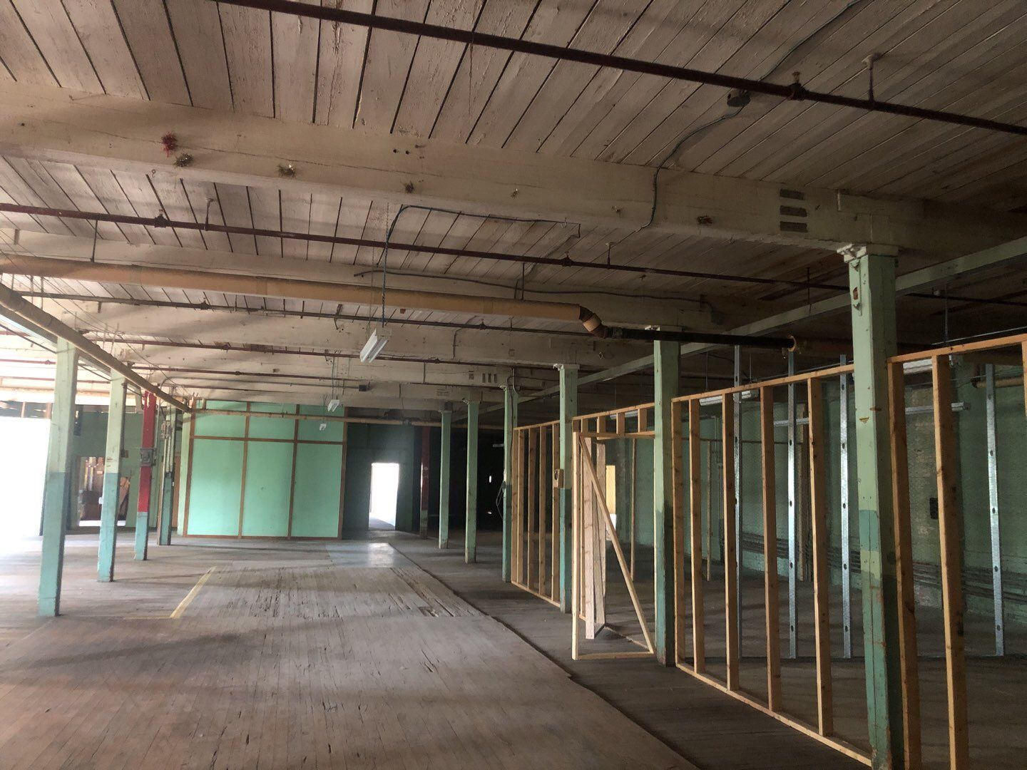 Interior view of a large, empty room in a building. Light-colored wooden floors, columns, and ceiling, with partially constructed walls on the right.