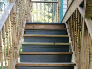 Wooden outdoor staircase leading upwards, painted gray treads, light wood railings with balusters, and partial view of greenery beyond.
