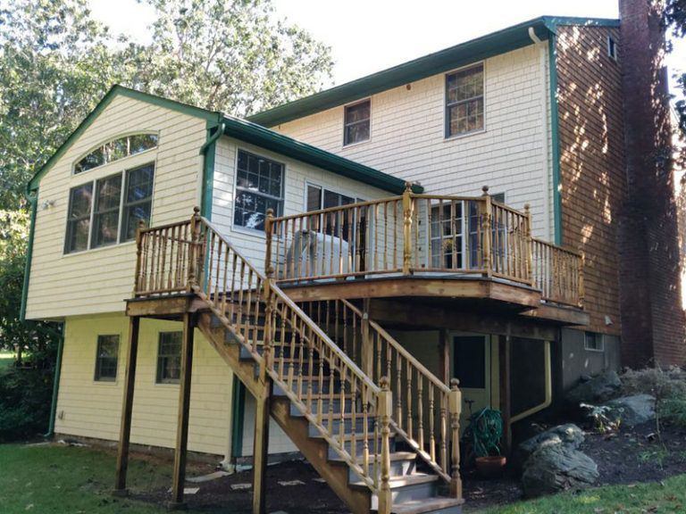 Two-story house with a wooden deck and staircase. The house has light siding, green trim, and a brick chimney.