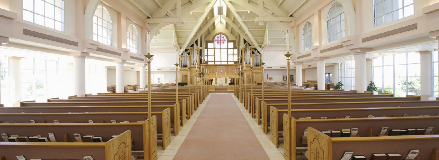 Interior view of a church with rows of wooden pews leading to the altar and a stained-glass window.