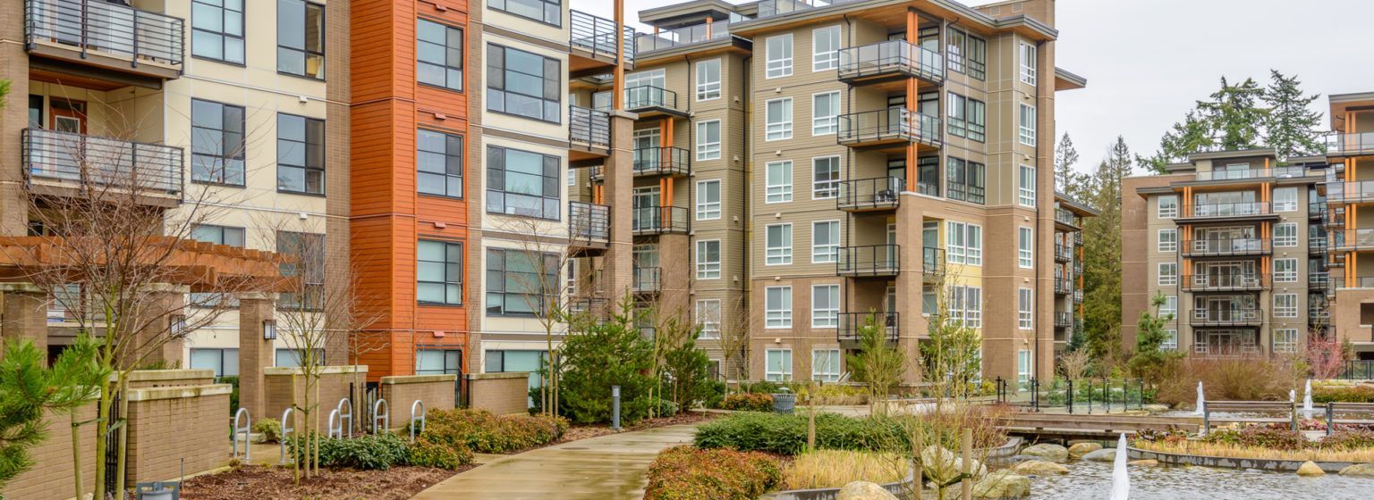 Apartment buildings with varying shades of brown and beige surrounded by landscaping and a water feature. Overcast day.