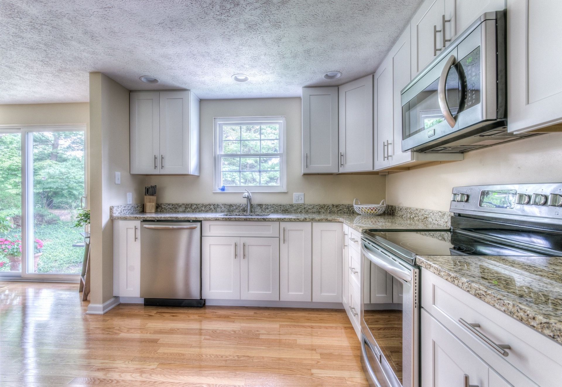 A bright kitchen with white cabinets, stainless steel appliances, granite countertops, and hardwood floors. A window and sliding glass door provide natural light.